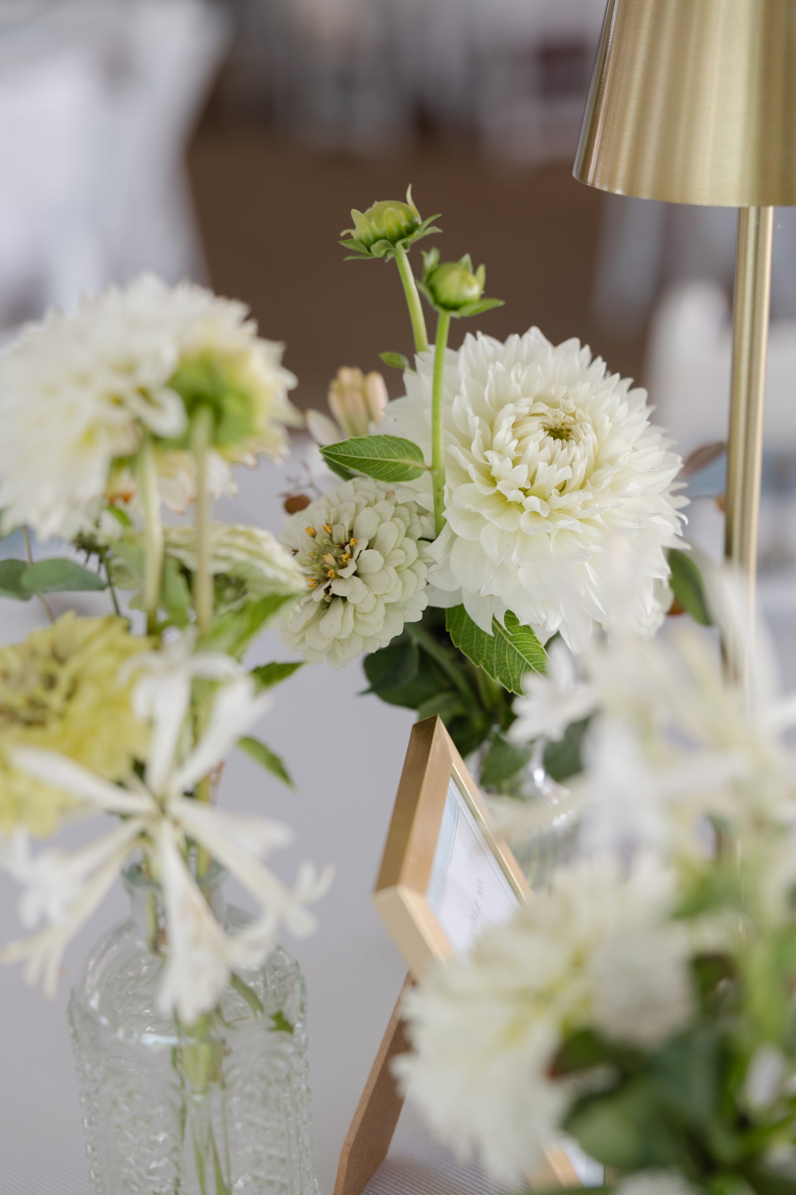 White dahlias and zinnias in glass vases beside a brass lamp on a reception table